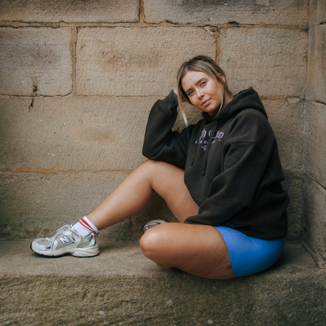 Woman sitting on stone steps wearing a brown tough cherry hoodie with combat essentials in pink text and blue shorts from tough cherry petal collection.