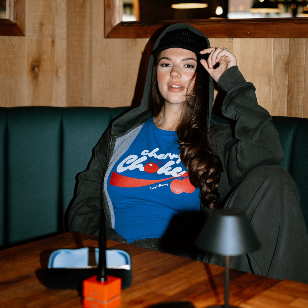 Woman wearing a grey hoodie with red and white tough cherry cherry choke top, sitting on a green couch in a room with wooden walls.