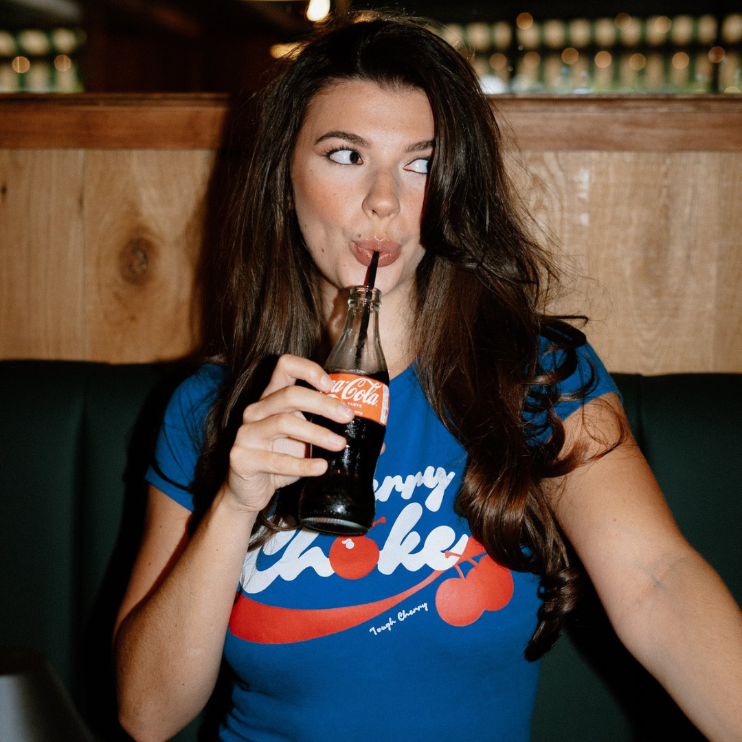 Woman drinking from a Coca-Cola bottle in a casual setting wearing tough cherry cherry choke top
