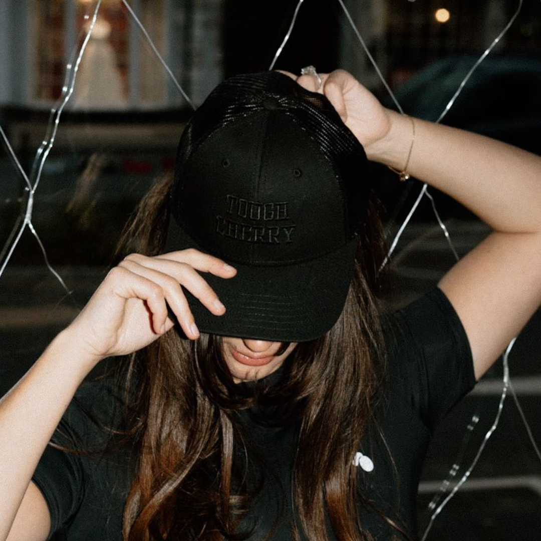Person wearing a black trucker cap with embroidered 'Tough Cherry', standing in front of a glass window.