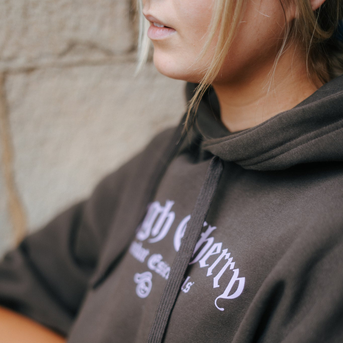 woman wearing a brown hoodie with pink combat essentials tough cherry text, standing against a neutral background