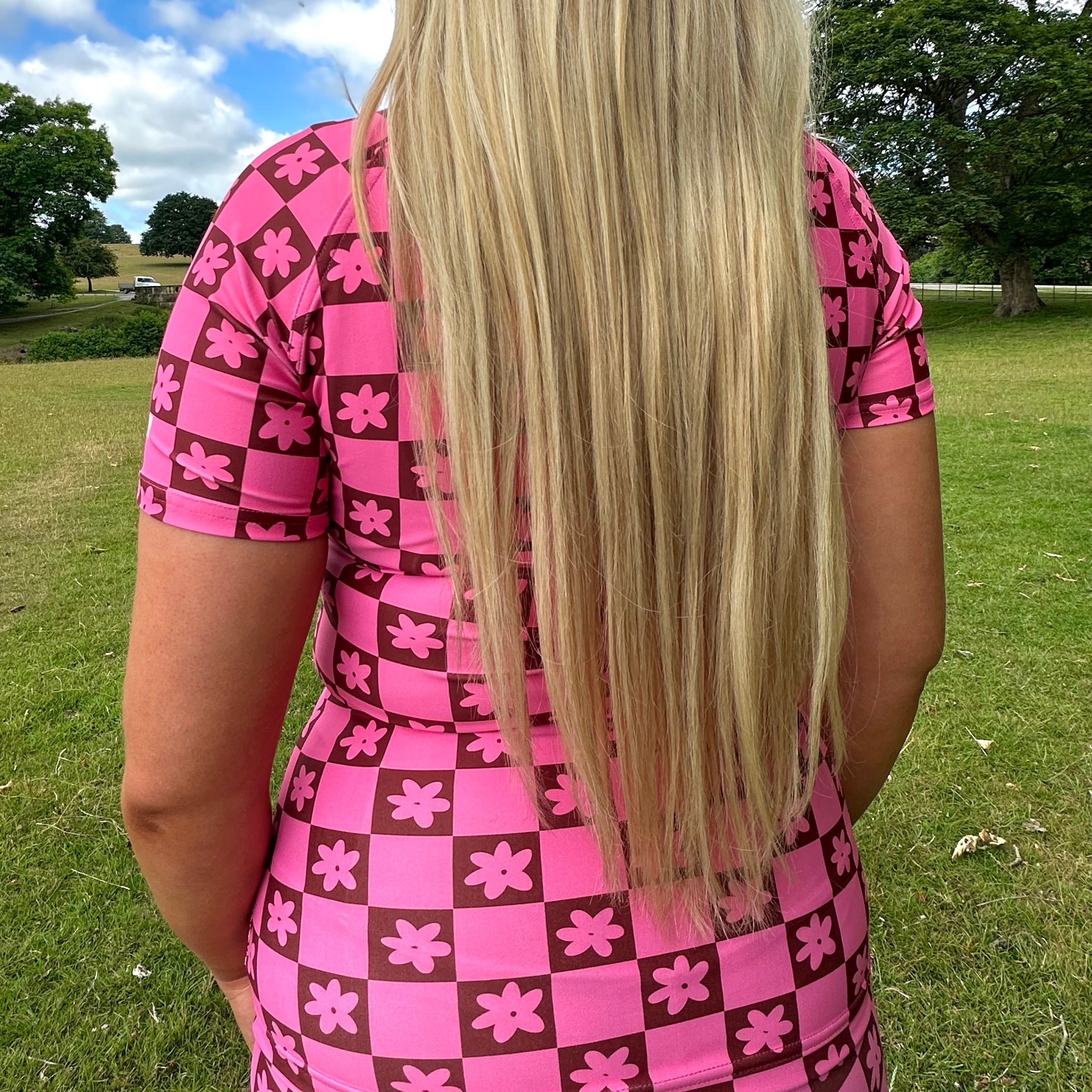 Woman wearing a Tough Cherry women's pink and Brown checkered jiu jitsu, grappling, no-gi Rash guard top with floral patterns in an outdoor setting back view.