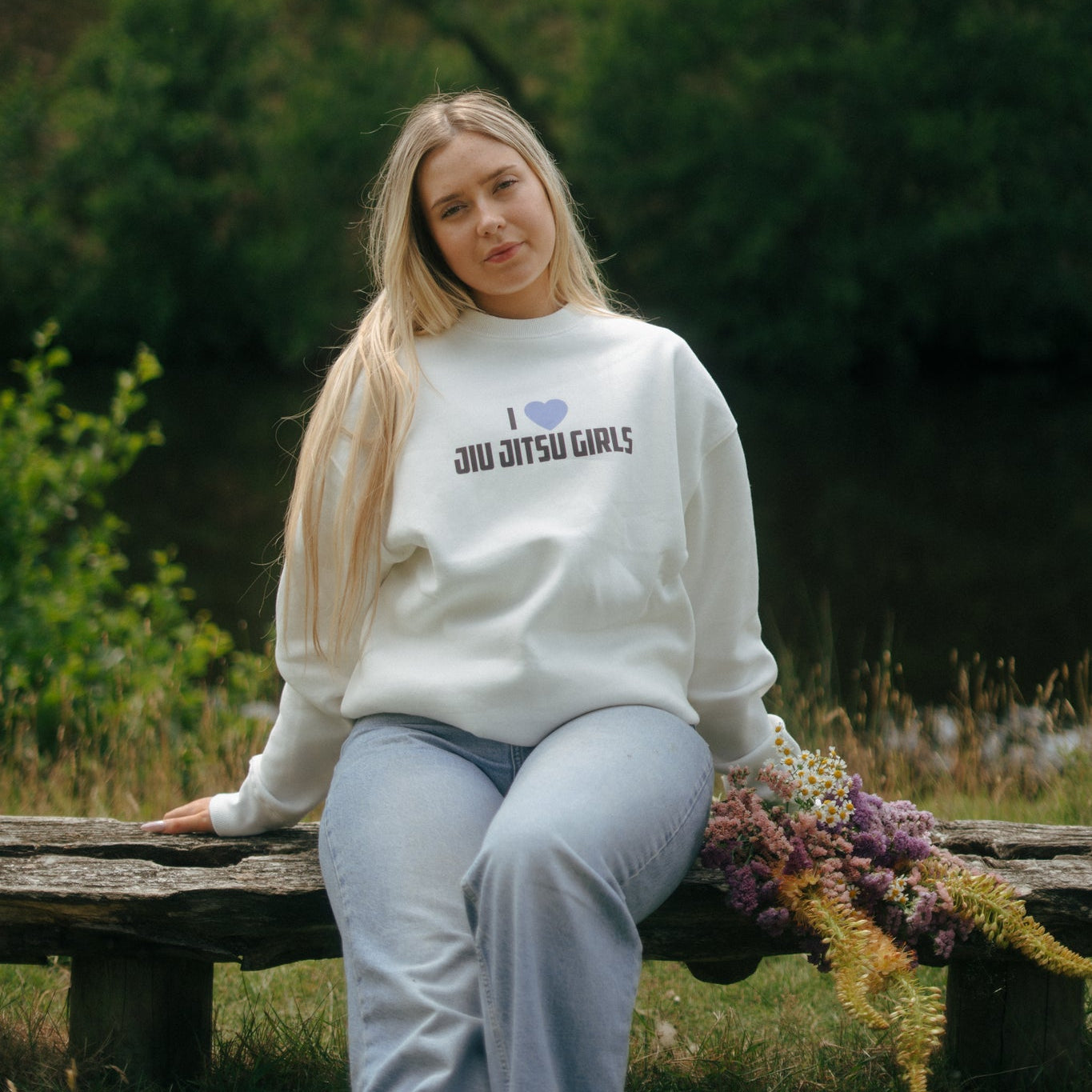 Woman wearing a white Tough Cherry sweatshirt with 'I love jiu jitsu girls' text, sitting on a wooden bench outdoors.
