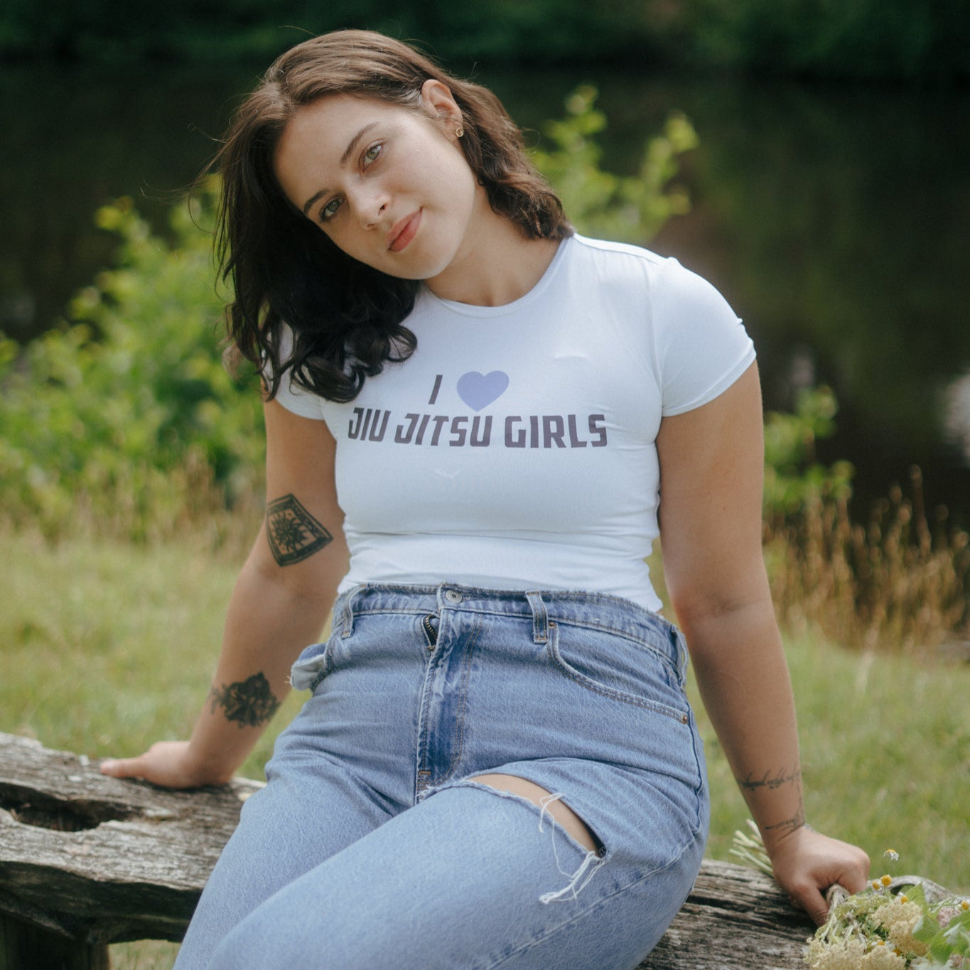 Woman sitting on a wooden bench outdoors wearing a white cropped Tough Cherry  t-shirt with text 'I love jiu jitsu girls' and blue jeans.