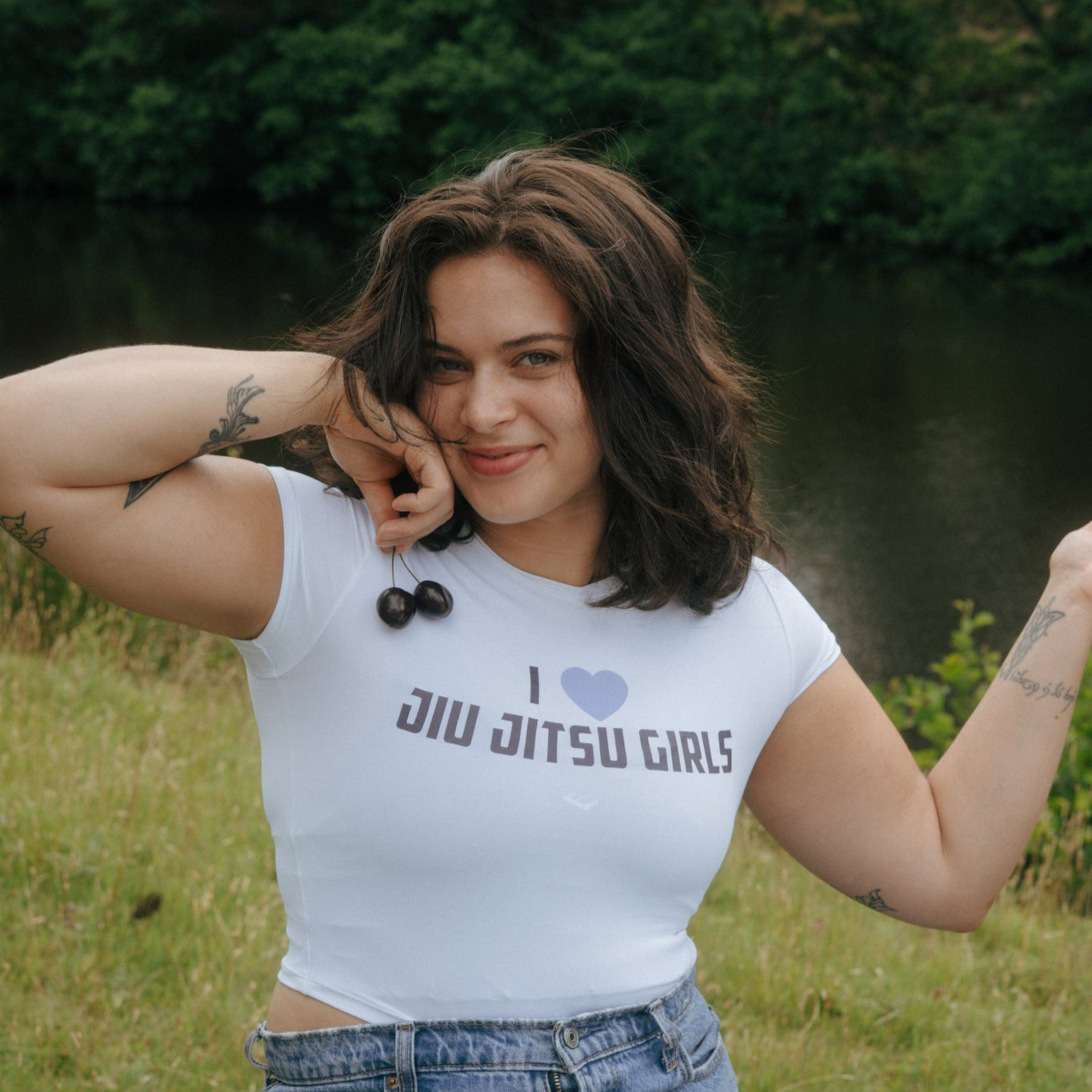 Woman wearing a 'I love Jiu Jitsu Girls' Tough Cherry cropped t-shirt sitting outdoors by a river holding cherries.