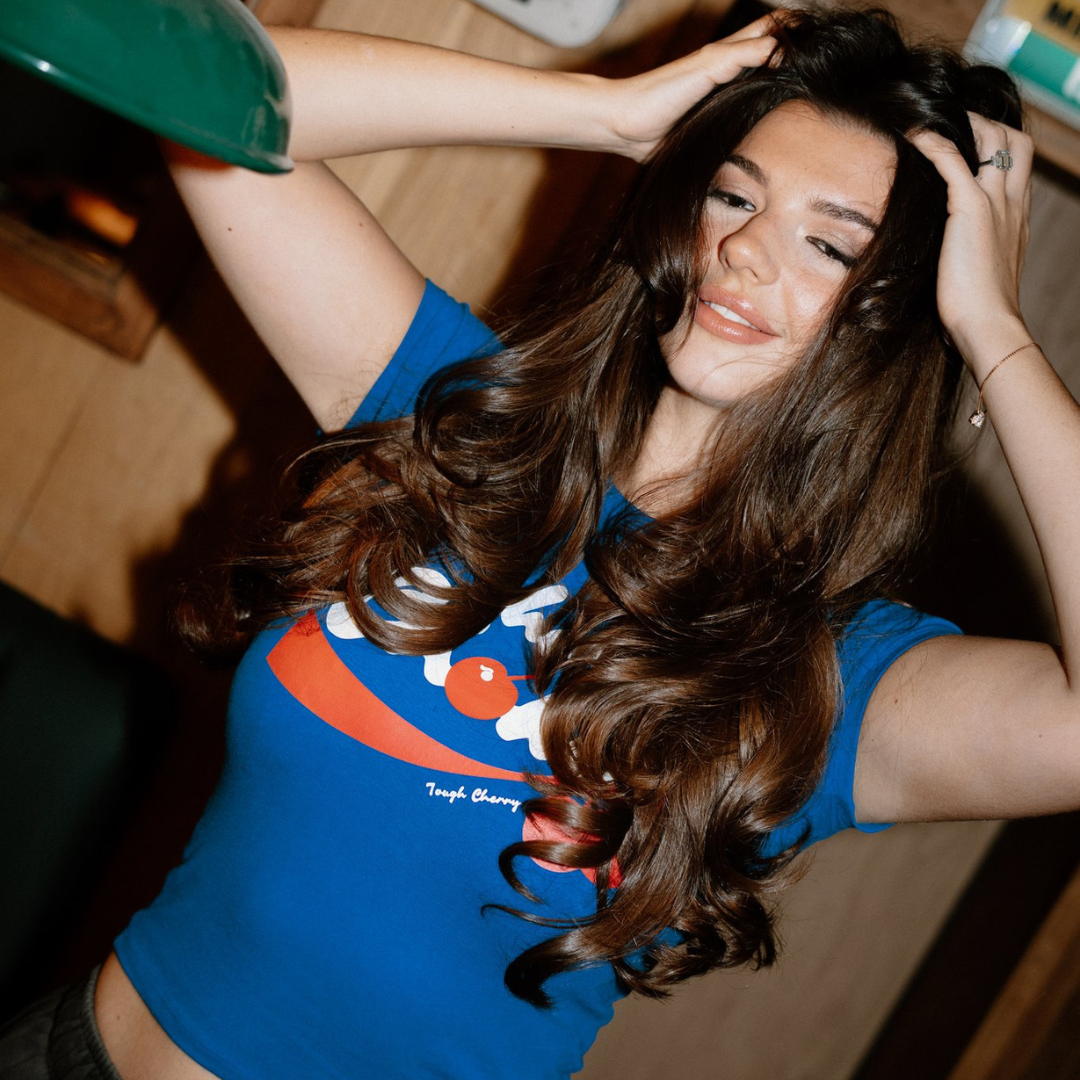 Woman with long brown hair wearing a blue shirt with a tough cherry cherry choke graphic.
