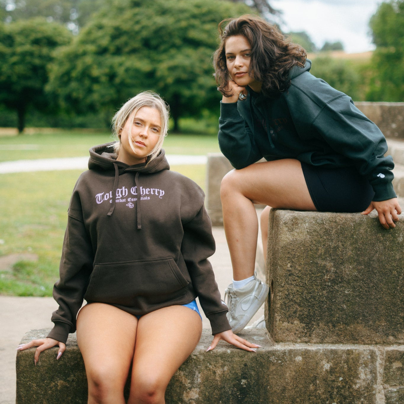 Two women wearing brown a green tough cherry hoodie for jiu jitsu sitting on stone steps outdoors with greenery in the background