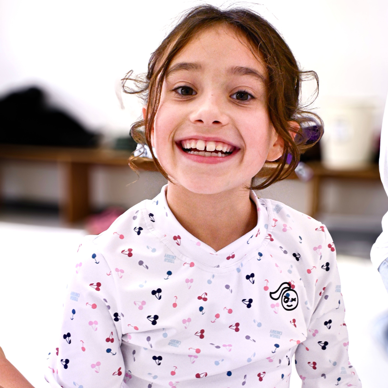 A girl wearing a tough cherry rash guard with cute cherries and a big smile with a blurred jiu jitsu gym background. 