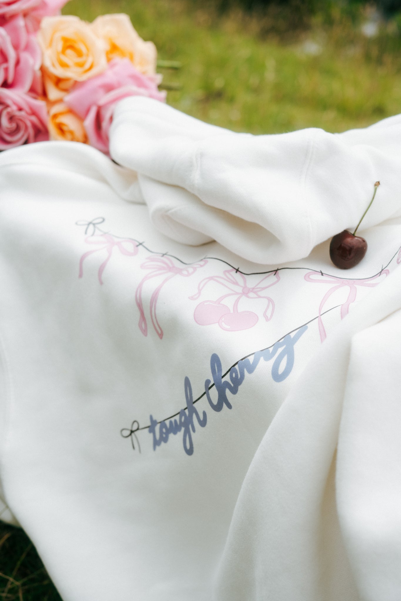 A woman wearing a white tough cherry hoodie with cherries and bows on it with a garden background. 