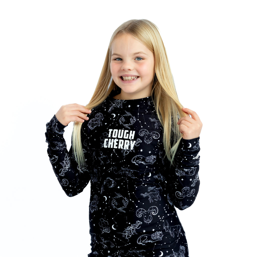 Children's under the stars long sleeved rash guard, black with white constellations print, blonde haired girl holding hair