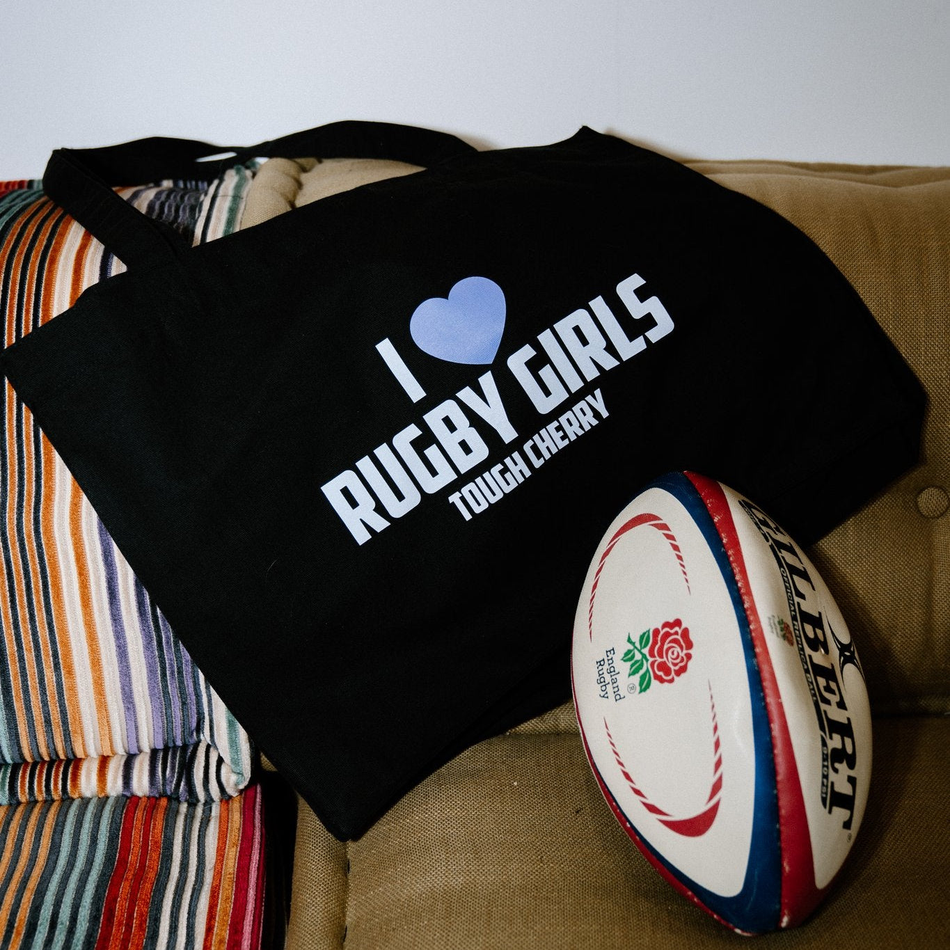 Black tote bag with 'I ❤️ Rugby Girls Tough Cherry' text on a couch next to a rugby ball.