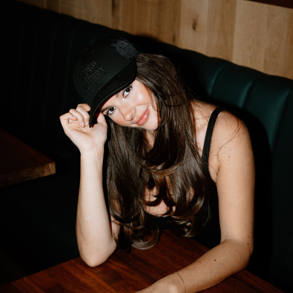Woman wearing a black tough cherry trucker cap sitting on a dark couch with a wooden table.