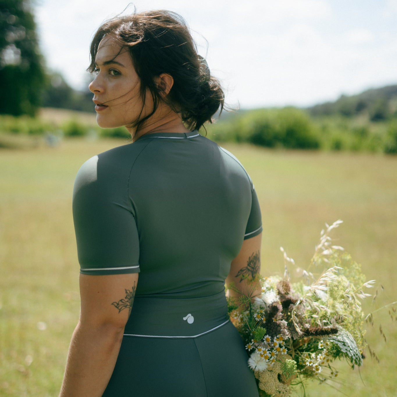Woman in green tough cherry rash guard and shorts standing in a field facing away holding a bunch of flowers