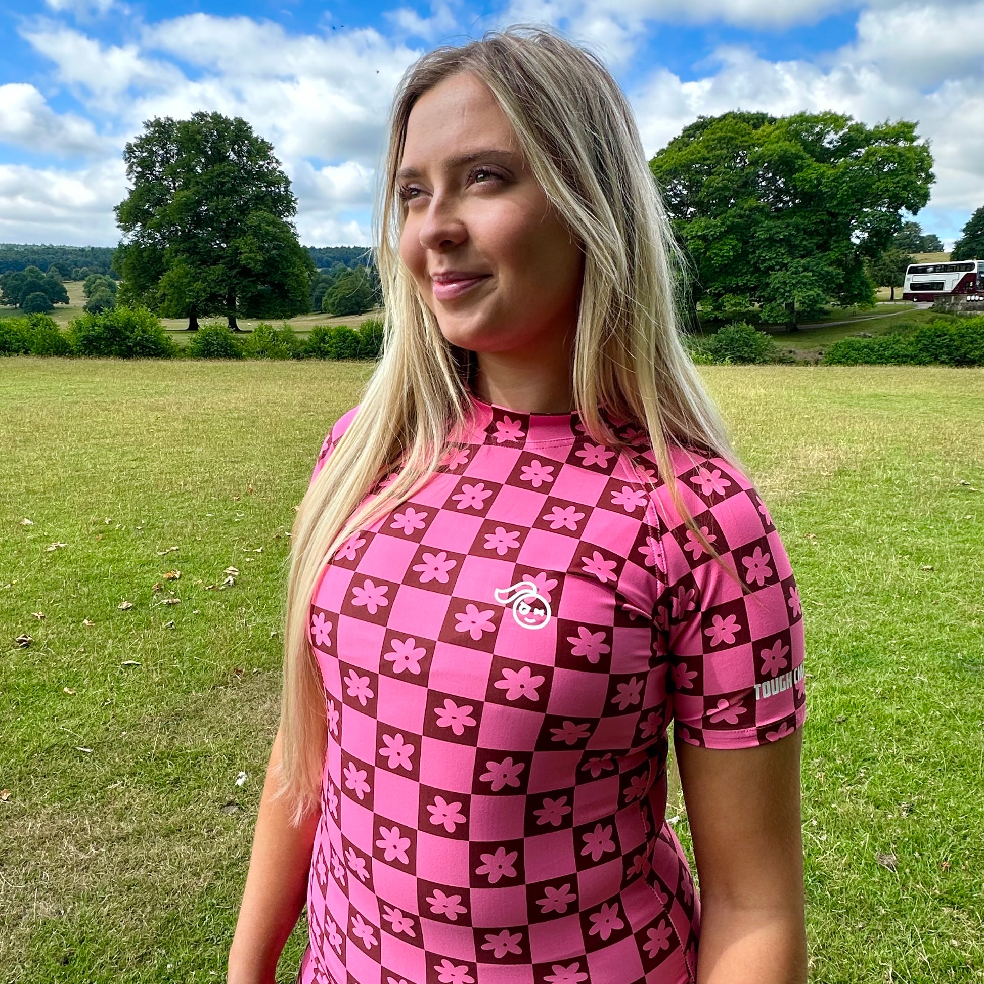 Woman wearing a Tough Cherry women's pink and Brown checkered jiu jitsu, grappling, no-gi Rash guard top with floral patterns in an outdoor setting. 