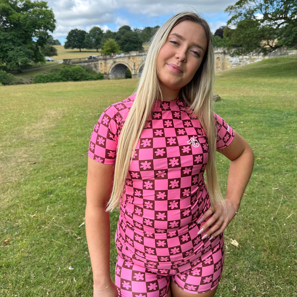 Woman wearing a Tough Cherry women's pink and Brown checkered jiu jitsu, grappling, no-gi Rash guard top with floral patterns in an outdoor setting full body view. 