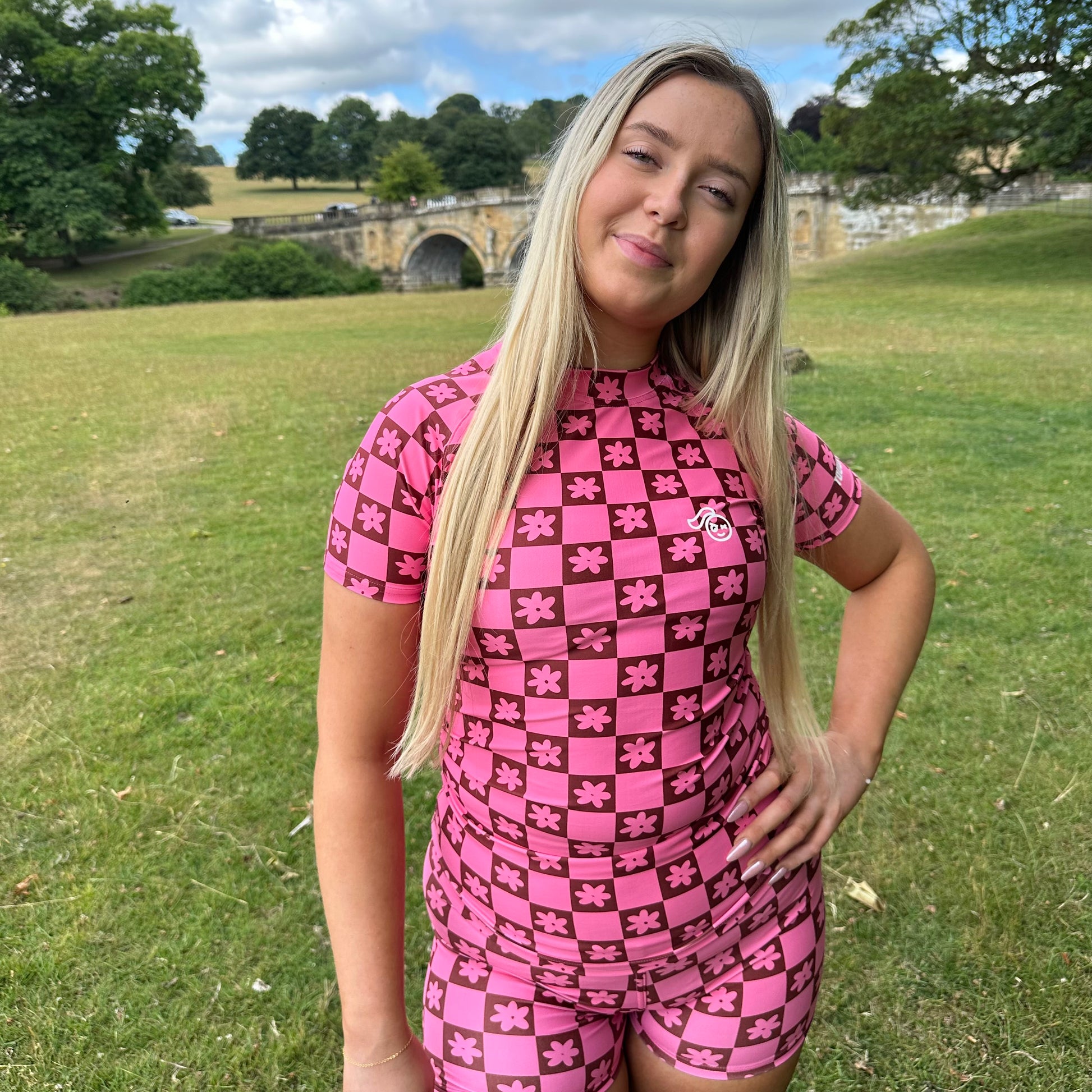 Woman wearing a Tough Cherry women's pink and Brown checkered jiu jitsu, grappling, no-gi Rash guard top with floral patterns in an outdoor setting full body view. 
