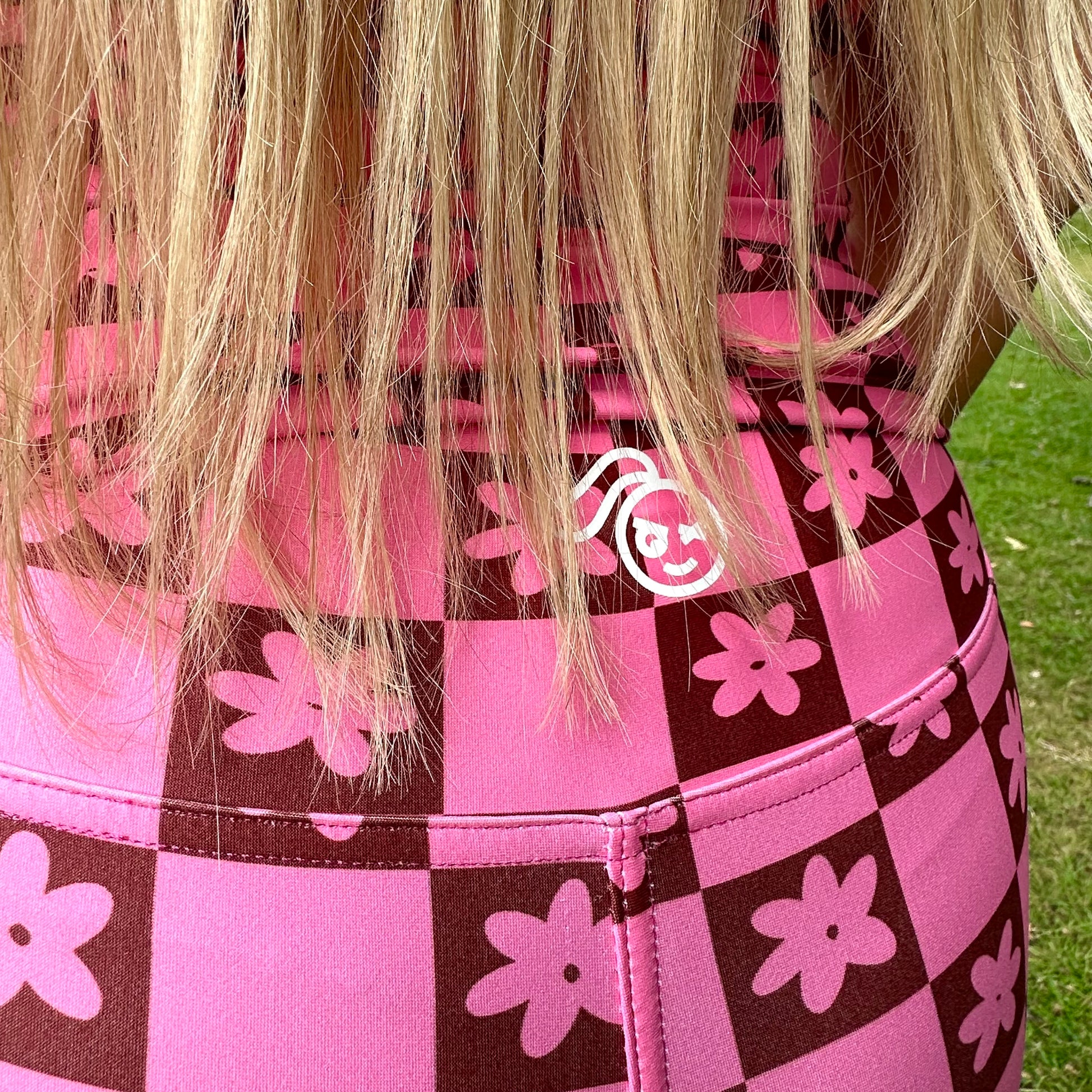 Woman wearing a Tough Cherry women's pink and Brown checkered jiu jitusu, grappling, no-gi high waisted shorts with floral patterns in an outdoor setting tough cherry logo view. 