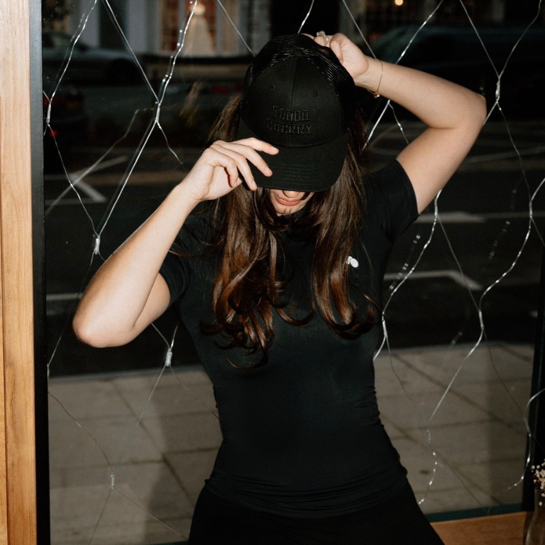 woman wearing a black tough cherry trucker cap and black tough cherry rash guard in front of a glass window.