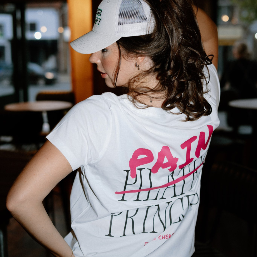 Person wearing a white t-shirt with pink and black text saying pain princess in an indoor setting