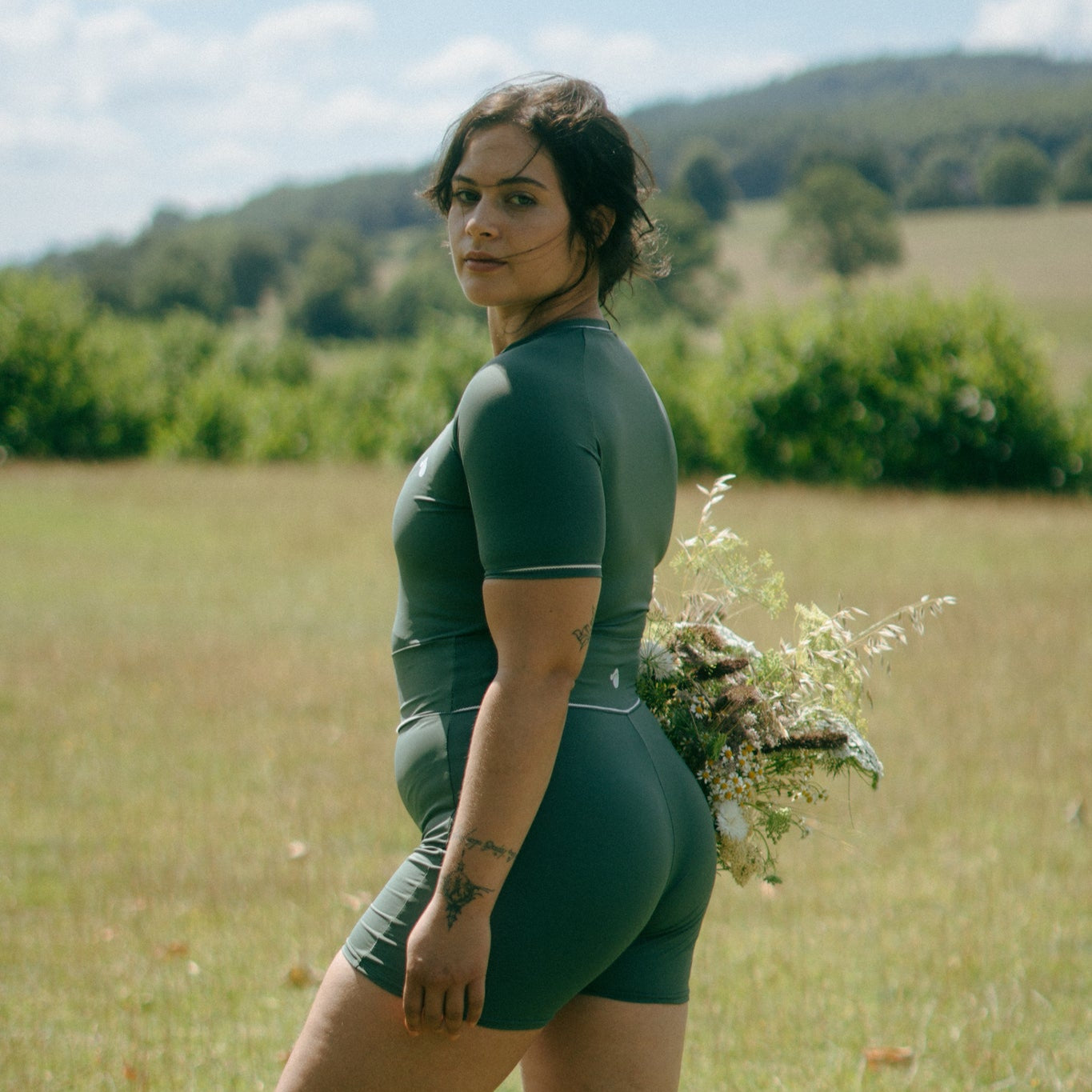 Woman in green tough cherry rash guard and shorts standing in a field with trees and blue sky in the background