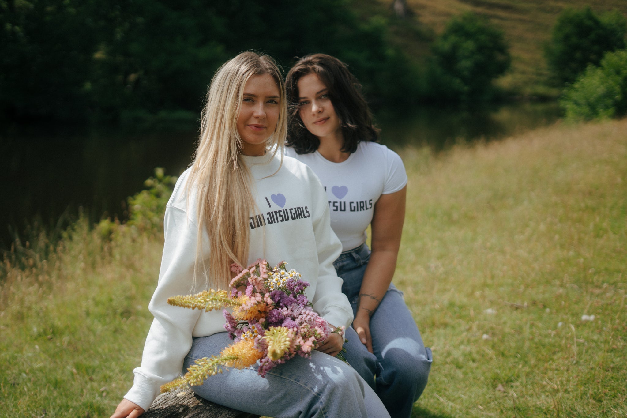 Two women sitting on a log in a field with flowers, wearing matching shirts from tough cherry with the text 'i love jiu jitsu girls'.