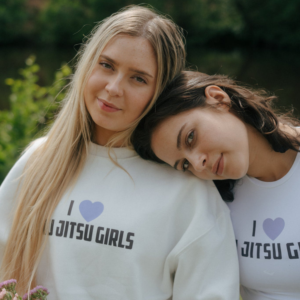 Two women wearing white tough cherry sweatshirts with 'I love Jiu Jitsu Girls' text, sitting outdoors.