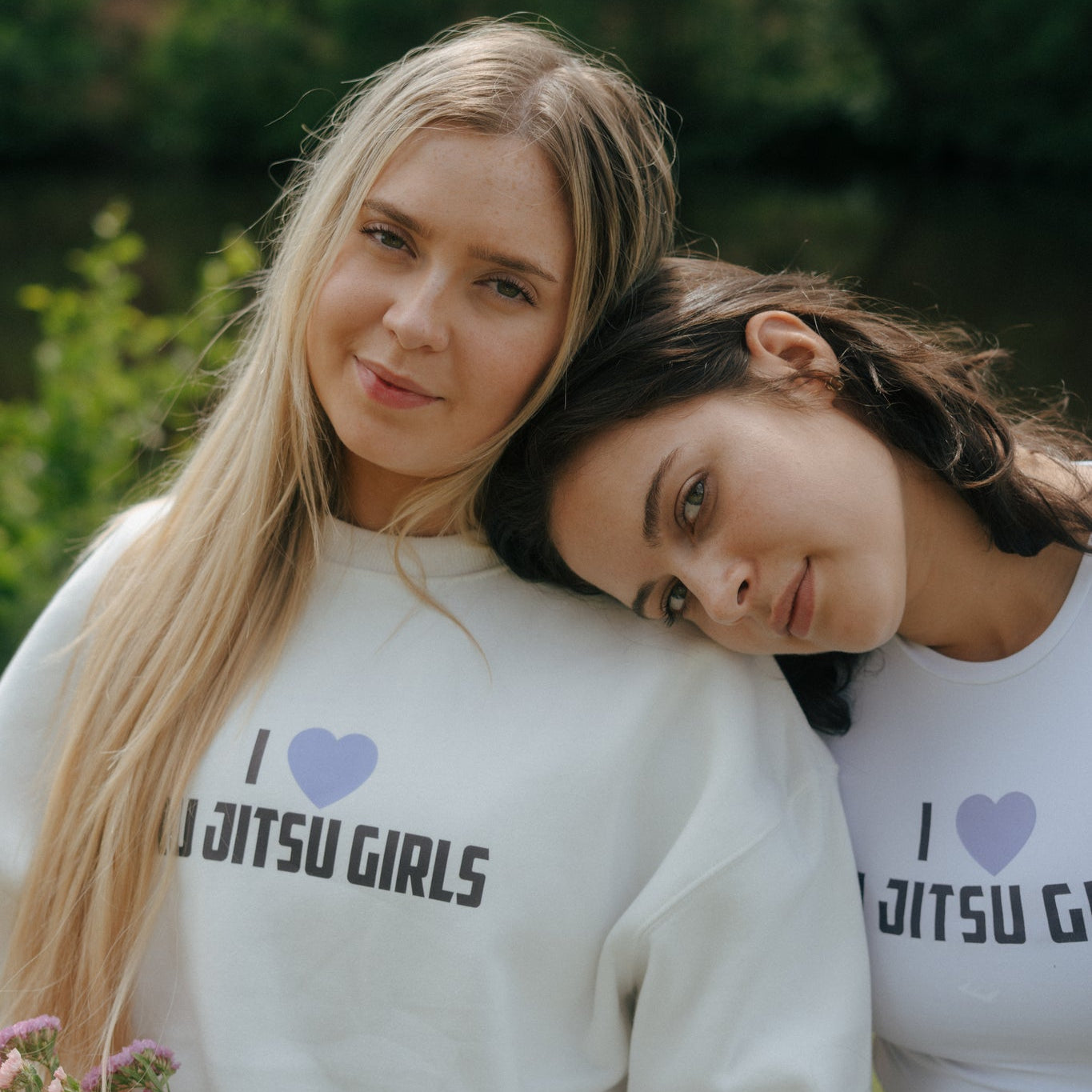 Two women wearing white tough cherry sweatshirts with 'I love Jiu Jitsu Girls' text, sitting outdoors.