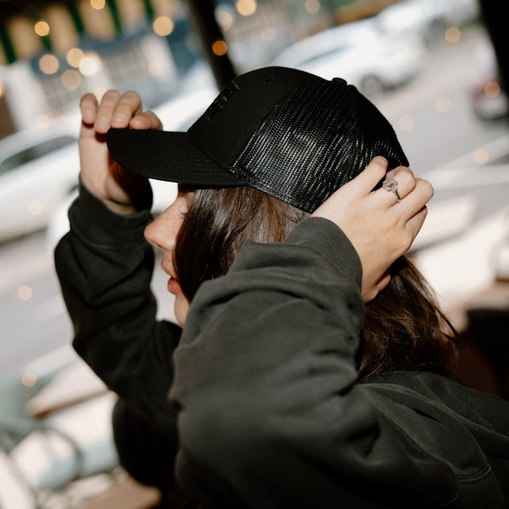 Person adjusting a black trucker cap indoors with blurred background
