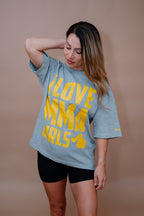 A woman wearing a heather gray oversized t-shirt with bold yellow text that reads 'I LOVE MMA GIRLS' and black athletic shorts, posing against a solid tan studio backdrop.