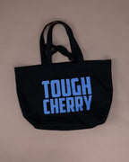 A black canvas tote bag with the words 'TOUGH CHERRY' printed in a bold light blue font on the front, displayed against a solid beige background.