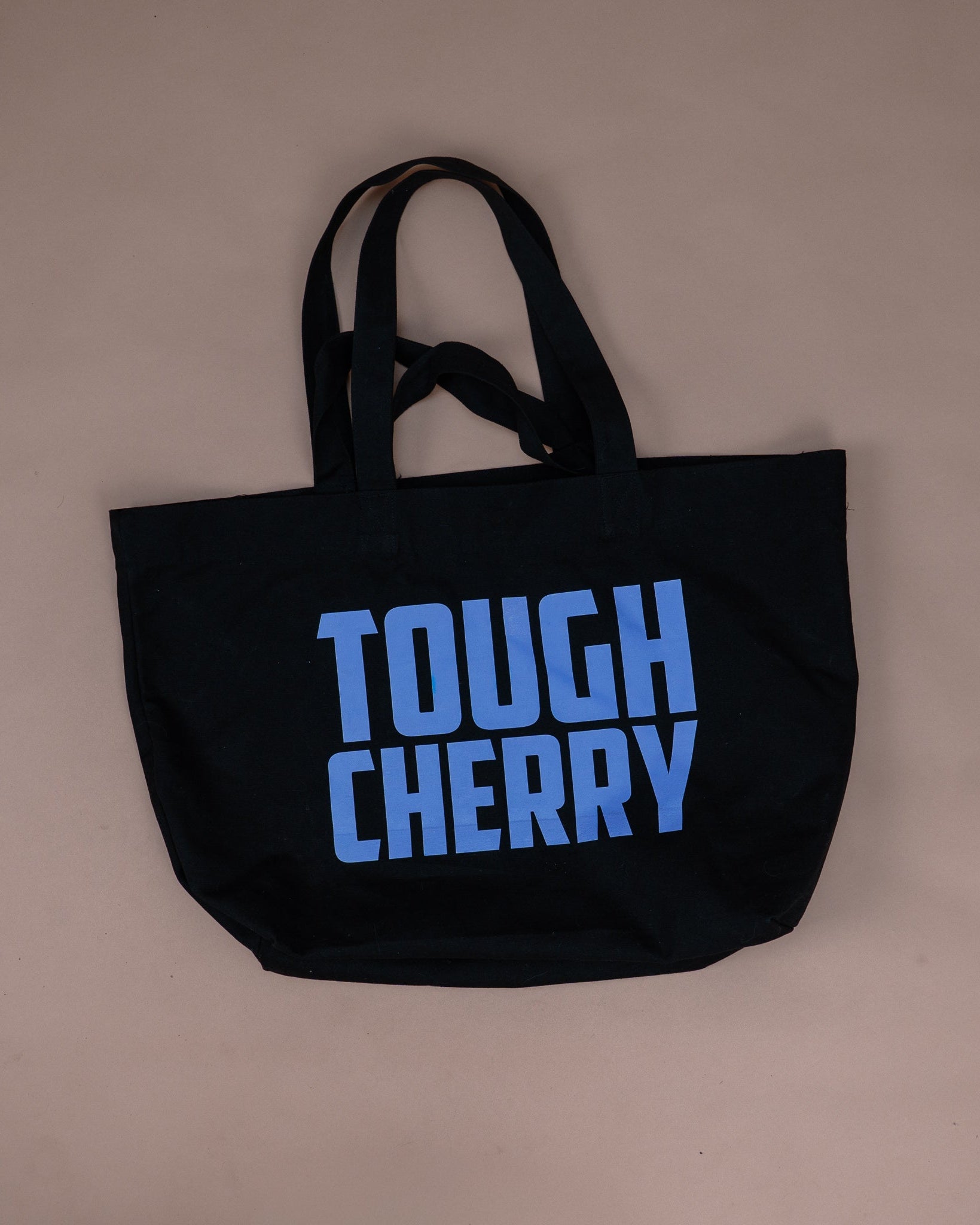 A black canvas tote bag with the words 'TOUGH CHERRY' printed in a bold light blue font on the front, displayed against a solid beige background.