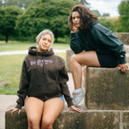 Two women wearing brown a green tough cherry hoodie for jiu jitsu sitting on stone steps outdoors with greenery in the background 